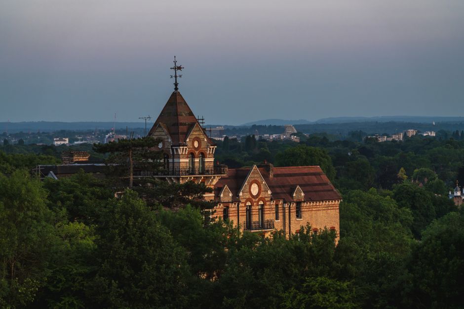 A historic brick church with a prominent triangular roof and a tall steeple topped with a weather vane, situated among dense green trees on a hillside. The church features arched windows and decorativ