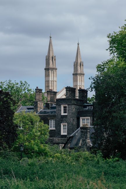 A historic brick church with a prominent triangular roof and a tall steeple topped with a weather vane, situated among dense green trees on a hillside. The church features arched windows and decorativ